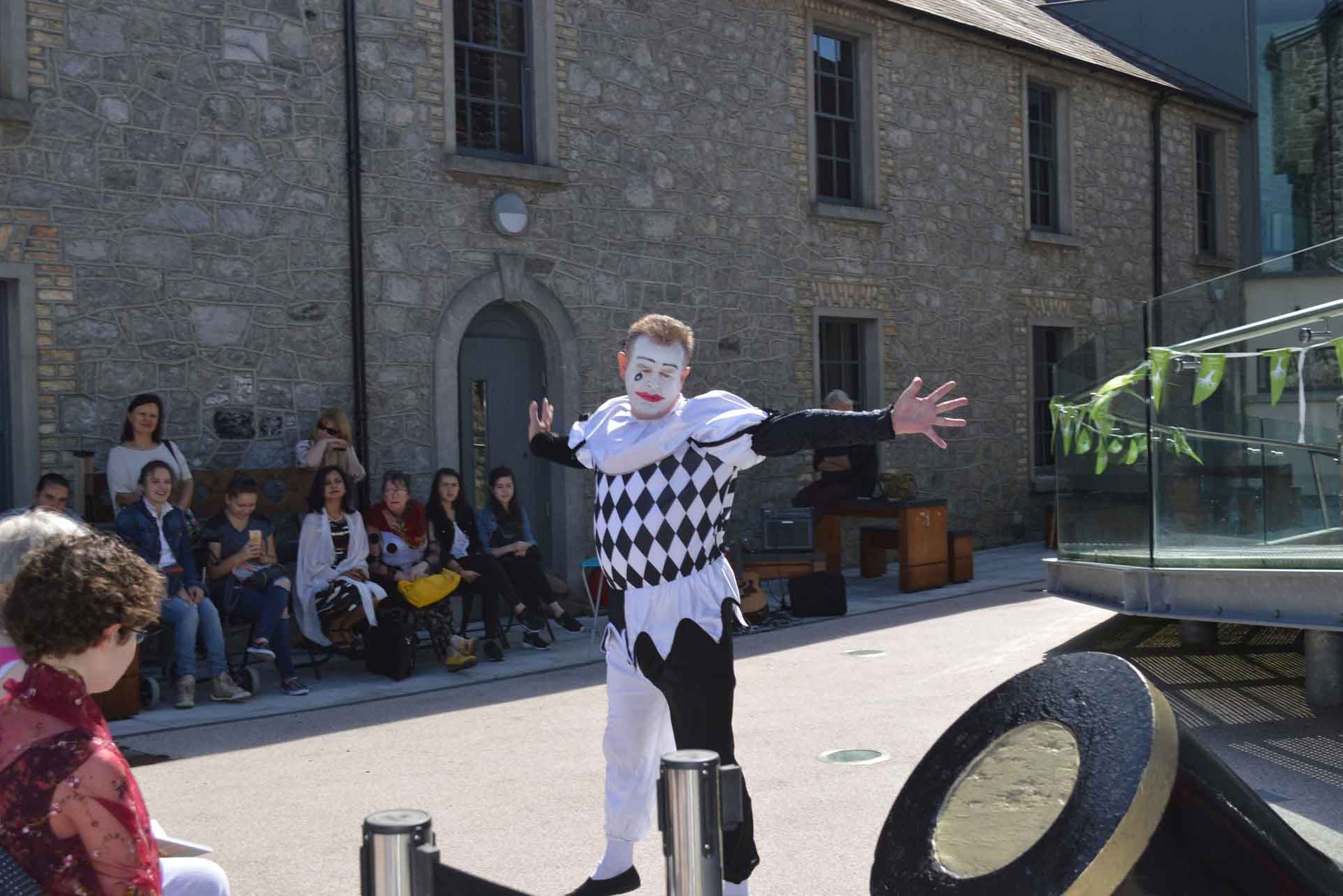 A crowd of visitors enjoying a special performance from a mime dressed in a black and white costume and face paint, as he entertains everyone with his act as part of our Poetry in the Park event at Athlone Castle courtyard.